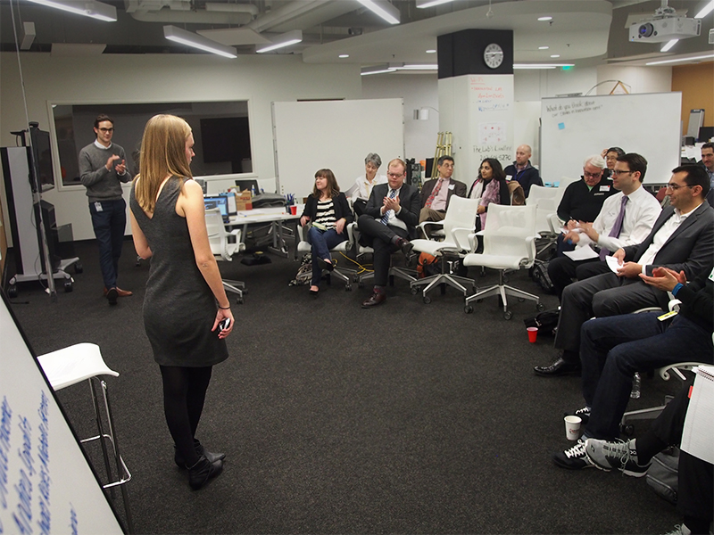 A photo showing a room with a grey carpet and white walls and whiteboards. The camera looks over the shoulder of a presenter with long blond hair, addressing a crowd of about 13 people in white rolling chairs.